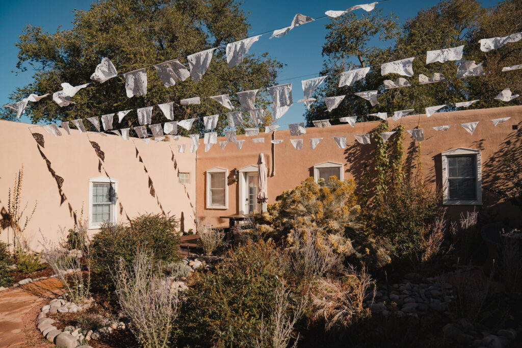 Wide courtyard view, papel picado strung overhead, wicker chairs and closed umbrella below